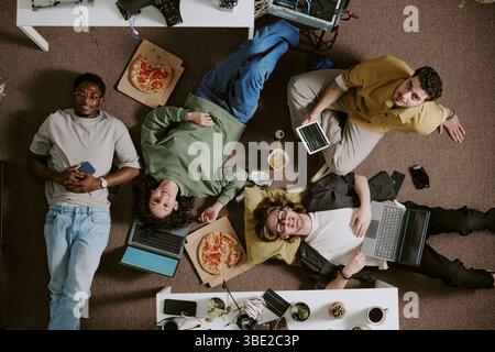 Eine Gruppe von Freunden genießt eine Pizzapause, während sie auf dem Boden mit Geräten und Gadgets zusammenarbeiten. Lässiges Ambiente mit Betonung von Teamwork und Kreativität Stockfoto
