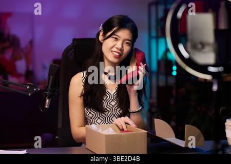 Frau lächelt und hält modischen roten Schuh, während sie Produkte im Studio überprüft. Hintergrund mit Mikrofon und Beleuchtung mit verschwommenem farbenfrohem Dekor Stockfoto