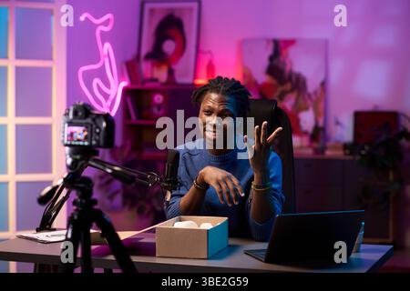 Filme von Content Creators in modernem, künstlerisch beleuchtetem Studio mit Laptop. Ansprechende Handgeste auf dem Schreibtisch mit Neonschild, das einen lebendigen Hintergrund reflektiert Stockfoto