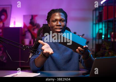 Porträt eines afroamerikanischen Mannes, der am Schreibtisch sitzt und eine Black Box mit Merchandise zur Online-Demonstration hält, in einem modernen, gut beleuchteten Studio mit violettem Neonlicht und Pflanzen Stockfoto