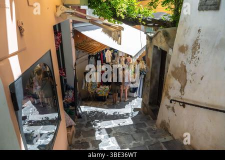 Positano, Italien an einem sonnigen Tag an der italienischen Amalfiküste stöbern Touristen in charmanten lokalen Geschäften in engen Gassen durch bunte Kleidung und Accessoires. Stockfoto