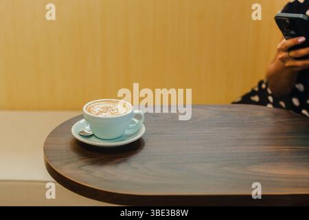 Tasse Cappuccino Kaffee auf Holztisch mit Frau mit Handy im Hintergrund. Stockfoto
