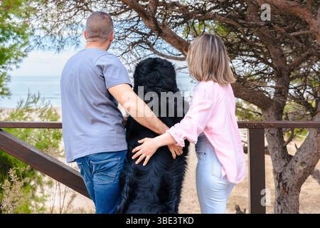 Ein Paar und ein großer schwarzer Hund mit Blick auf das Meer von einer Holzterrasse in der Natur Stockfoto