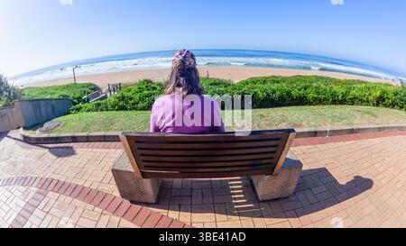 Frau, die auf der Promenade Bank sitzt, mit Blick auf den Strand, das blaue Meer und die Landschaft des Himmels, eine Nahaufnahme hinten hinter dem Foto. Stockfoto