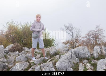 Eine Frau mittleren Alters lächelt auf einem nebeligen Bergpfad, der von felsigen Pfaden und Herbstbäumen umgeben ist und leichte Sportbekleidung trägt. Stockfoto