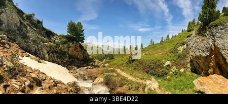 Erkunden Sie die friedliche Landschaft einer alpinen Region mit grünen Tälern, fließenden Bächen, Felsen und verschneiten Flecken unter einem ruhigen blauen Himmel, ideal für Stockfoto