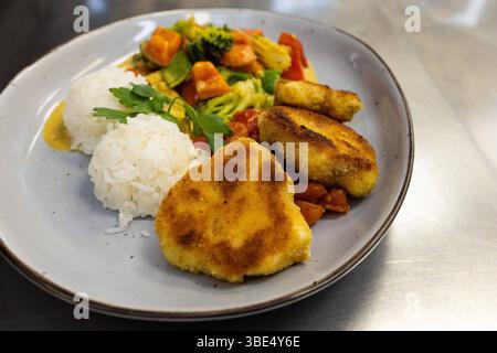 Deutschland 07. April 2025: Im Bild: Eine appetitliche Portion goldbraun panierter Blumenkohlschnitzel, serviert mit Lockerem weißem Reis und einer reichhaltigen Mischung aus gedünstetem Gemüse, darunter Brokkoli, Karotten, Paprika und Zucchini. Das vegetarische Gericht ist ansprechend auf einem grauen Teller arrangiert und mit frischer Petersilie garniert. Ein Hauch von Kremiger Soße umspielt die Komponenten. Dieses Foto eignet sich hervorragend vegetarische Ernährung, fleischlose Gerichte, gesunde Rezepte, Mittagessen, Abendessen, Familienmahlzeiten, Catering, Kantinenessen, Schulessen, pfla Stockfoto