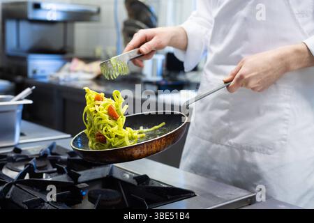 Deutschland 07. April 2025: Im Bild: Ein Koch in weißer Arbeitskleidung schwenkt gekonnt Pasta mit getrockneten Tomaten und grüner Soße in einer rustikalen Pfanne über einen Gasherd in einer professionellen Restaurantküche. Die Bewegung des Schwenkens ist dynamisch eingefangen, wobei die Nudeln in der Luft sind und das können des Kochs unterstreichen. Der Hintergrund zeigt eine typische, moderne Küchenumgebung mit Edelstahloberflächen und professionellen Geräten. Dieses Bild eignet sich ideal für Kochen, Gastronomie, Restaurantbetrieb, Küchenpersonal, Kochkünste, Zubereitung von Speisen, itali Stockfoto