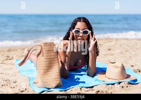 Eine junge Frau entspannt sich am Strand, trägt eine stilvolle Sonnenbrille und trägt eine Strandtasche Stockfoto