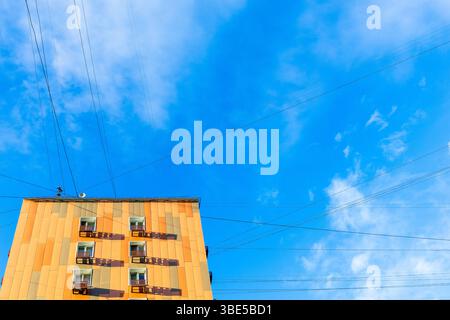 Ansicht von unten auf ein mehrstöckiges Wohngebäude, das mit orangefarbenen Seitenverkleidungen und elektrischen Leitungen und Kabeln im blauen Himmel bedeckt ist Stockfoto