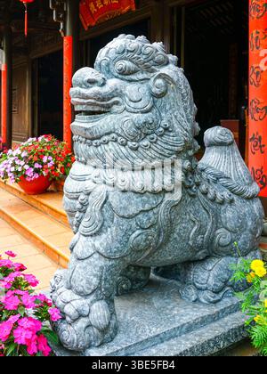 Detaillierter Blick auf die traditionelle Steinhüter-Löwe-Statue vor der vietnamesischen Tempeltreppe mit bunten Blumen und roten Säulen Stockfoto
