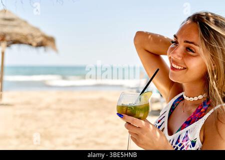 An einem warmen Sommernachmittag entspannt sich eine Frau am Strand und genießt ein buntes Getränk, während sie die Sonne genießt. Die Meereswellen schaffen eine ruhige Kulisse Stockfoto