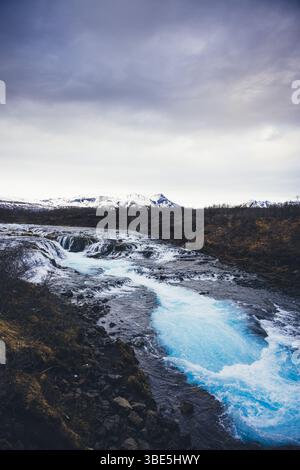 Der atemberaubende Bruarfoss-Wasserfall, bekannt für sein kristallklares türkisfarbenes Wasser, fließt über dunkle vulkanische Felsen, umgeben von Islands zerklüfteter Landschaft Stockfoto