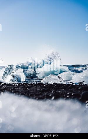 Fesselnde Eisformationen am Diamond Beach, Island, Kontrast zum schwarzen Sand, mit Wellen, die dynamisch im Hintergrund unter klarem Licht zusammenbrechen Stockfoto