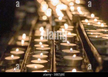 Eine Reihe von kleinen, brennenden Kerzen in Metallhaltern, die ein warmes und gemütliches Licht erzeugen, vielleicht in einer Kirche oder einem anderen heiligen Ort. Stockfoto