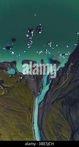 Vertikale Drohnenaufnahme eines geflochtenen Gletscherflusses, der auf eine grüne Lagune trifft, mit Eisbergen, die in der Nähe des Ufers schwimmen. Eine lebendige Luftaufnahme von Islands roher gla Stockfoto