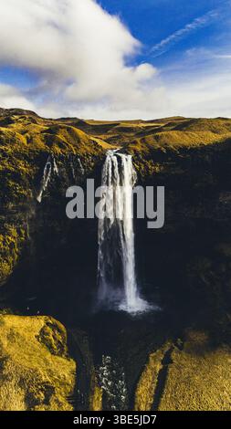 Atemberaubende Aussicht auf den Seljalandsfoss-Wasserfall in Island, der von einer hohen Klippe umgeben von goldenen Hügeln unter einem leuchtend blauen Himmel mit sanftem Himmel herabstürzt Stockfoto