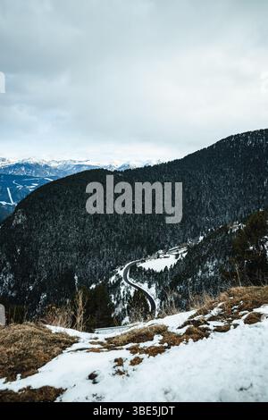 Eine gewundene Straße schlängelt sich durch eine verschneite Berglandschaft in Andorra. Der bewölkte Himmel sorgt für einen kühlen Ton und verbessert die dramatische Sicht der immergrünen Abdeckung Stockfoto