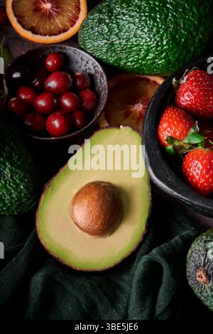Eine frische Auswahl an Früchten, darunter Avocado, Erdbeeren, Preiselbeeren und Orangenscheiben auf dunklem Hintergrund, die ein lebendiges und Stockfoto
