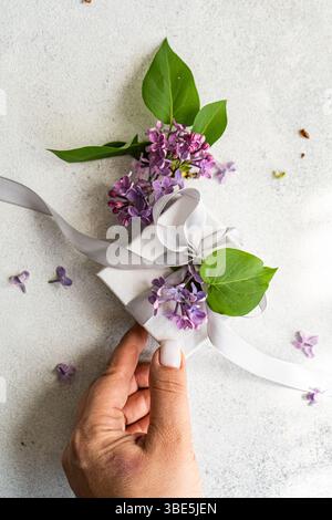 Eine zarte Geschenkbox mit lilafarbenen Blumen und grünen Blättern, gebunden mit einem weichen weißen Band. Eine Hand präsentiert anmutig das elegante Paket auf einem li Stockfoto