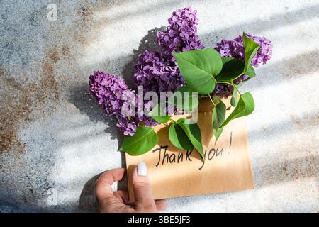 Eine Hand hält eine Karte mit einem Dankeschön! Nachricht mit leuchtenden lila Blumen auf strukturiertem Hintergrund. Sonnenlicht wirft sanfte Schatten über t Stockfoto