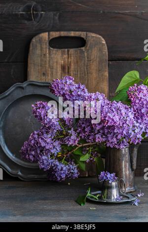 Charmante Anordnung von Fliederblüten in einer Metallvase gegen rustikales Holz. Die leuchtenden violetten Farbtöne verleihen der einfachen Komposition einen Hauch von Vintage-Eleganz Stockfoto