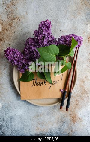 Ein brauner Umschlag mit Dankeschreiben, verziert mit leuchtenden lila Blumen und grünen Blättern. Holzstäbchen ruhen auf einer neutralen Platte. Stockfoto
