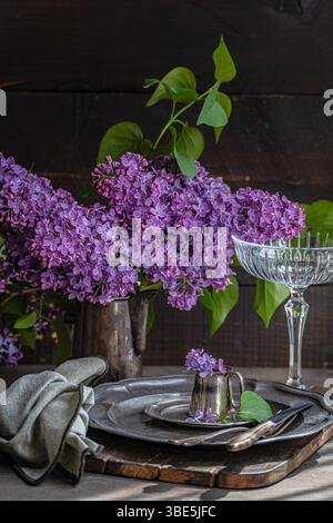 Ein eleganter Tisch mit leuchtenden Fliederblüten in einem rustikalen Krug. Vintage-Besteck und Glaswaren ergänzen das florale Arrangement auf einem Holz Stockfoto