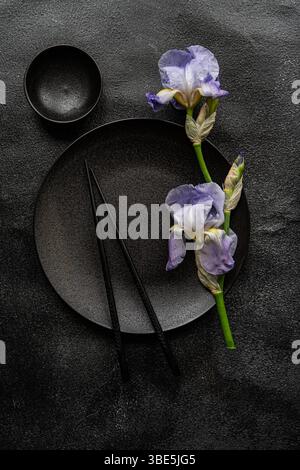 Draufsicht auf elegante Irisblumen neben schwarzem Keramikgeschirr auf einem strukturierten dunklen Hintergrund, der einen auffälligen visuellen Kontrast erzeugt, der s betont Stockfoto