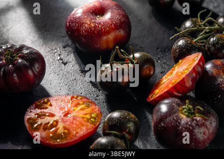 Eine Auswahl verschiedener Erbstücke, einschließlich halbierter und ganzer Tomaten, glitzernd mit Wassertropfen, auf dunkler Oberfläche für einen rustikalen Bio-Look Stockfoto