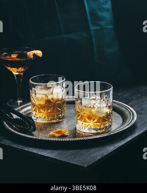 Zwei mit Eis gefüllte Whiskey-Gläser stehen auf einem silbernen Tablett zusammen mit einem Cocktail mit leuchtender orangener Dekoration auf einem dunklen und eleganten Rücken Stockfoto
