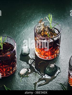 Eine dramatische Szene, in der Whiskey in einem mit Rosmarin garnierten Glas spritzt, umgeben von verstreuten Eiswürfeln auf einer dunklen, strukturierten Oberfläche, die ein Moo erzeugt Stockfoto