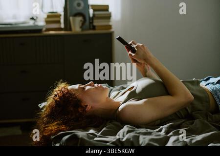 Frau mit rotem Haar, die sich auf dem Bett zurücklehnt, vertieft darin, ihr Mobiltelefon in gemütlicher Atmosphäre zu überprüfen. Bücher und helle Dekorationsstücke im Hintergrund Stockfoto