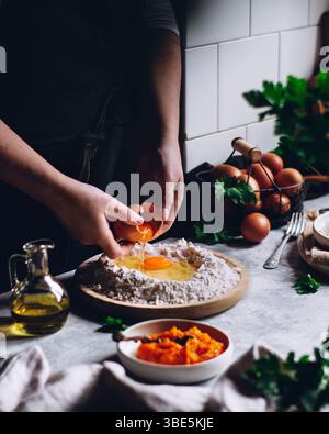 Eine Person zerbricht ein Ei in einen Mehlhügel, umgeben von einer rustikalen Küche mit Kräutern, frischen Eiern und Olivenöl, wodurch ein warmes, hausgemachtes entsteht Stockfoto