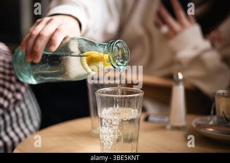 Das zugeschnittene, nicht erkennbare Mädchen gießt Wasser aus einer Flasche mit einem Stück Zitrone in ein Glas in einem Café und fängt einen Moment der Erfrischung ein. Stockfoto
