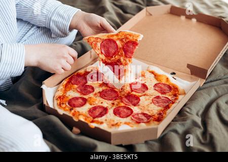 Eine nicht wiedererkennbare Frau, die gemütlich ein Stück Peperoni-Pizza isst, während sie bequem in ihrem Bett sitzt, während die Pizzabox neben ihr liegt. Stockfoto