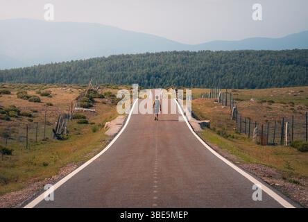 Ein einsamer Mann geht eine lange, leere Straße entlang, die von Zäunen gesäumt ist und von Feldern umgeben ist, und geht in Richtung der fernen Hügel unter klarem Himmel in Guadarrama Mo Stockfoto