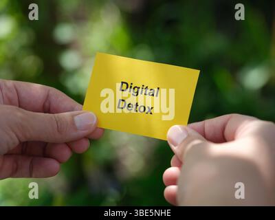Eine Frau, die eine gelbe Papiernote mit den Worten Digital Detox in den Händen hält, vor einem Laubhintergrund. Nahaufnahme. Stockfoto