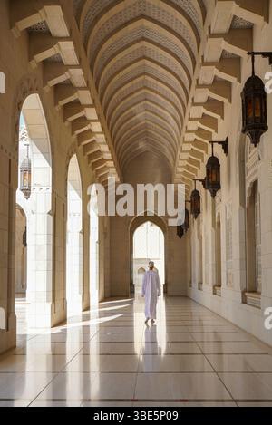 Rückansicht eines nicht erkennbaren Mannes in traditioneller Dschdasha-omanischer Kleidung, der entlang eines Korridors in der Sultan-Qaboos-Moschee in Maskat, Oman, spaziert Stockfoto