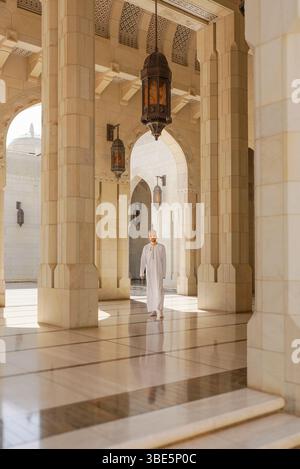 Ein Mann in traditioneller omanischer Dschasha-Kleidung spaziert durch den großen Korridor der Sultan-Qaboos-Moschee in Muscat, eingerahmt von kunstvollen Bögen und hängenden La Stockfoto