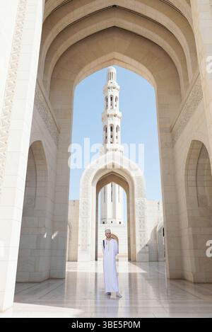 Ein Mann in traditioneller omanischer Kleidung steht unter einem Bogen in der Sultan-Qaboos-Moschee in Maskat, Oman, mit einem detaillierten Minarett im Hintergrund. Stockfoto