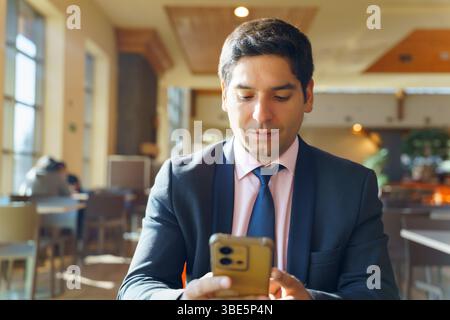 Junger chilenischer Geschäftsmann in einem modernen Café, im Anzug und mit einem Smartphone. Sonnenlicht filtert durch große Fenster und schafft so einen Beruf Stockfoto