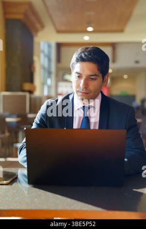 Ein junger chilenischer Geschäftsmann in formeller Kleidung arbeitet fleißig an seinem Laptop in einem modernen Café. Sonnenlicht filtert durch große Fenster und schafft so einen Fokus Stockfoto