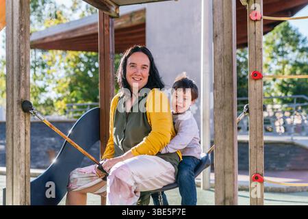 Eine fröhliche Mutter und ein Kind mit Down-Syndrom sitzen an einem sonnigen Tag zusammen auf einer Spielplatzschaukel. Das Bild fängt einen warmen und glücklichen Moment mit ein Stockfoto