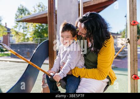 Eine fröhliche Mutter und ihr lächelnder Sohn mit Down-Syndrom teilen sich einen Moment auf einer Schaukel auf einem sonnigen Spielplatz. Die Szene fängt Wärme, Liebe und Spaß in einem ein Stockfoto