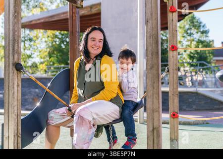 Eine Mutter und ihr Kind mit Down-Syndrom teilen einen freudigen Moment auf einer Schaukel auf dem Spielplatz. Die Szene fängt die Wärme der Familienbindung in der Sonne ein Stockfoto