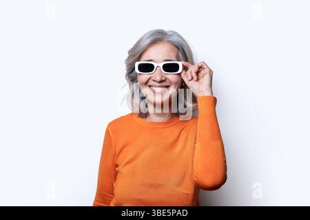 Eine asiatische chinesische Seniorin mit grauem Haar, die gerne eine stilvolle weiße Sonnenbrille und einen orangefarbenen Pullover trägt und ein Gefühl von Selbstvertrauen und Freude ausstrahlt Stockfoto