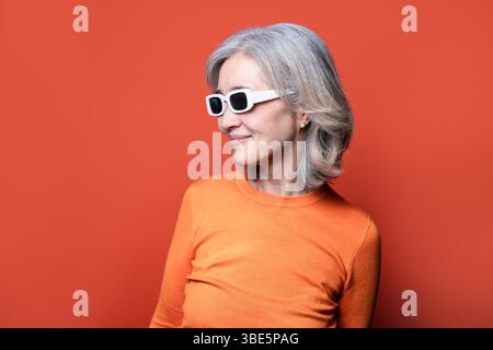 Asiatische chinesische Seniorin mit grauem Haar, trendige weiße Sonnenbrille und orangefarbenem Pullover. Sie steht vor einem orangefarbenen Hintergrund und verkörpert Co Stockfoto
