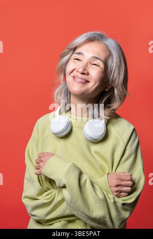 Asiatische Chinesische Seniorin trägt ein helles Sweatshirt und Kopfhörer, lächelt warm vor einem leuchtend roten Hintergrund, verkörpert Freude und Entspannung Stockfoto