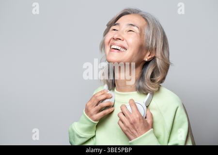 Asiatische Chinesische Seniorin lächelt fröhlich, während sie Kopfhörer hält und ihre Lieblingsmusik genießt. Das Bild fängt Glück und Freizeit in einer entspannten Atmosphäre ein Stockfoto